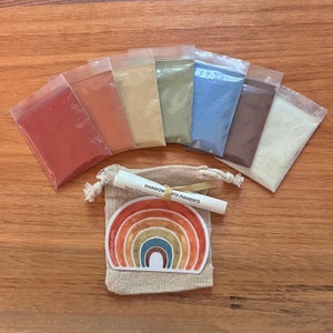Puede incluir: Un conjunto de siete peque&ntilde;as bolsas que contienen pigmentos de tierra de diferentes colores. Las bolsas est&aacute;n dispuestas en fila sobre una superficie de madera. Una peque&ntilde;a bolsa con un dise&ntilde;o de arco&iacute;ris y el texto "Pigmentos de tierra arco&iacute;ris" est&aacute; en el centro de la imagen.