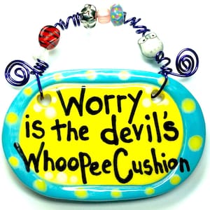May include: A blue and yellow ceramic sign with white polka dots. The sign reads "Worry is the devil's whoopee cushion".
