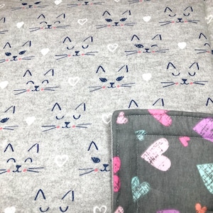 May include: Gray fabric with a repeating pattern of cat faces and white hearts. A corner of a darker gray fabric with pink, purple, and teal hearts is visible. The cat faces have navy blue eyes and whiskers.