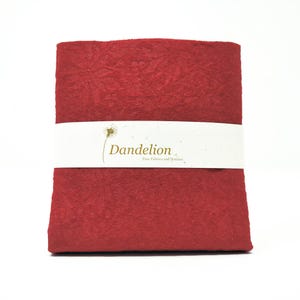 Peut inclure: Un paquet plié de tissu rouge, maintenu par une bande blanche. La bande porte le mot "Dandelion" en écriture élégante, et la légende "Fine Fabrics and Notions". Le tissu a un aspect texturé, probablement un textile.