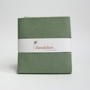Op de afbeelding: Een stofstaal van saliegroen vilt met een wit label waarop "Dandelion Fine Fabrics and Notions" staat.