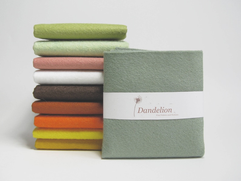 May include: A stack of 10 felt fabric squares in various colors, including green, pink, brown, orange, and yellow. The top square is a solid sage green with the text "Dandelion" printed on a white label.