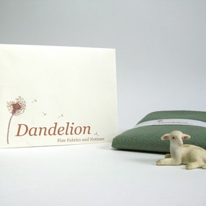 May include: A white envelope with a brown dandelion graphic and the text "Dandelion Fine Fabrics and Notions". A small green fabric square with the word "Dandelion" printed on it and a white lamb figurine are in the foreground.