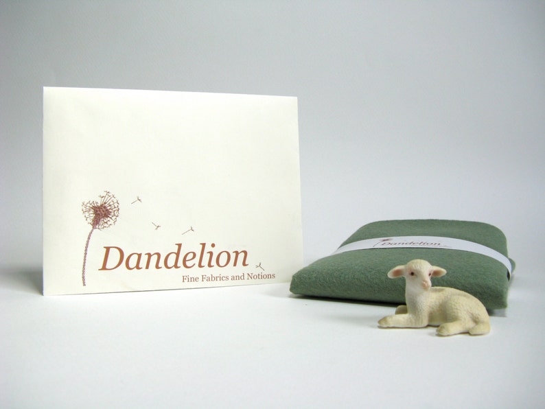 May include: A white envelope with a brown dandelion graphic and the text "Dandelion Fine Fabrics and Notions". A small green fabric square with the word "Dandelion" printed on it and a white lamb figurine are in the foreground.