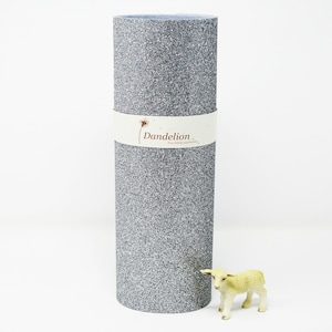 May include: A roll of silver glitter fabric with the text "Dandelion" on a white label. The fabric is rolled up and standing on a white surface. A small white toy lamb is in the foreground.