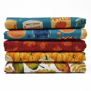 Könnte beinhalten: Ein Stapel gefalteter Stoffe mit herbstlichen Mustern. Die Stoffe zeigen Designs von Äpfeln, Kürbissen und Herbstblättern in Orange-, Gelb-, Rot- und Türkistönen. Ein Stoff hat den Text "Sweet Jessie's Louisiana".