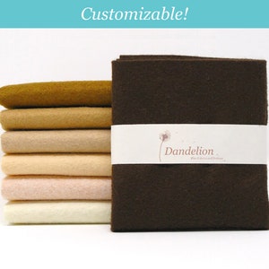 May include: A selection of felt fabric in shades of brown, beige, and cream. A dark brown felt bundle is wrapped with a white band displaying the "Dandelion" logo and the text "Fine Fabrics and Notions". The word "Customizable!" is written in the top left corner.