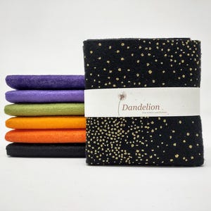 Peut inclure: Une pile de carrés de tissu en feutre colorés en violet, lavande, vert, orange et noir. Une plus grande pile de tissu noir avec des mouchetures dorées est enveloppée d'une bande blanche avec le texte "Dandelion" en marron.