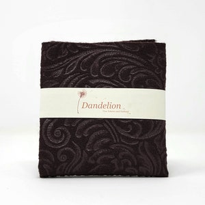 Könnte beinhalten: Ein braunes Stoffmuster mit einem geschwungenen, geprägten Muster. Der Stoff ist mit "Dandelion Fine Fabrics and Notions" beschriftet.
