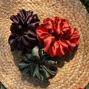 May include: Four satin scrunchies in shades of burgundy, rust, and olive green, arranged on a woven surface. The scrunchies are bunched and gathered, showcasing their shiny texture. These hair accessories add a touch of elegance to any hairstyle.
