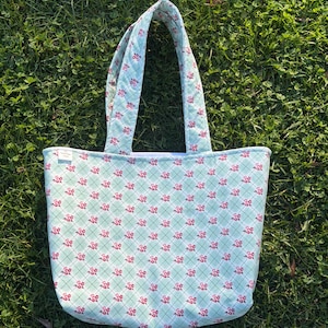 May include: A quilted tote bag with a light blue background and a repeating pattern of small red flowers. The bag has matching handles and a rectangular shape, perfect for carrying items.