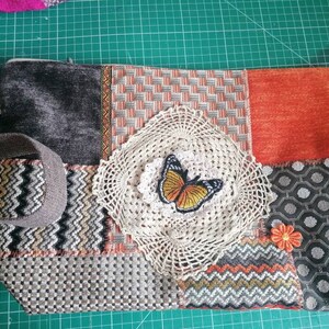 May include: A patchwork fabric bag with a zipper closure, a brown strap, and a crocheted doily with an orange butterfly applique. The bag is made of different fabrics in shades of gray, orange, and brown.