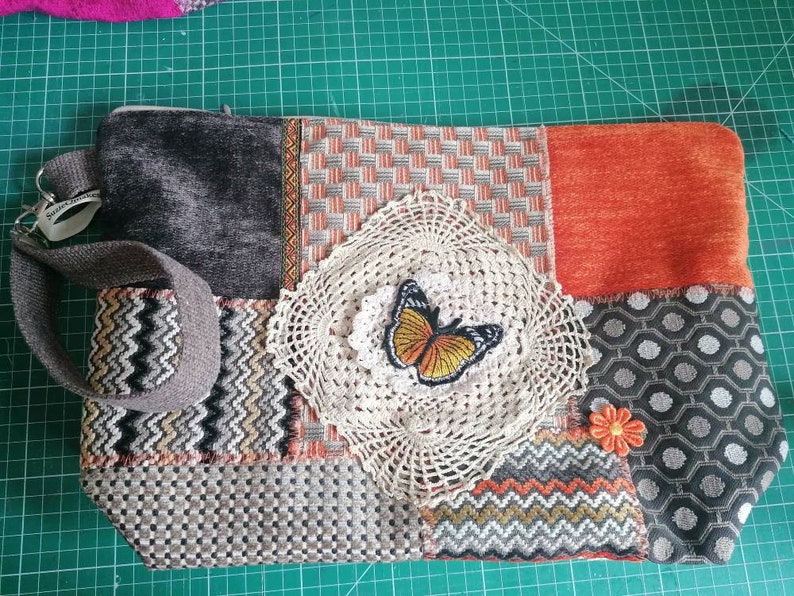 May include: A patchwork fabric bag with a zipper closure, a brown strap, and a crocheted doily with an orange butterfly applique. The bag is made of different fabrics in shades of gray, orange, and brown.