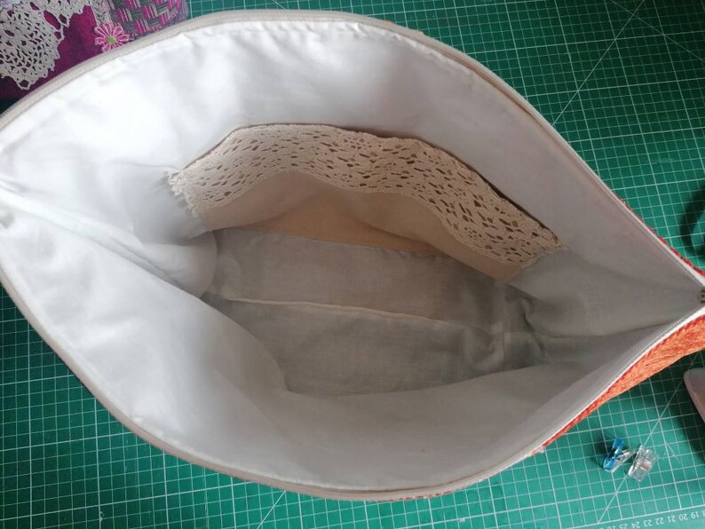 May include: The inside of a red and white zippered pouch with a white lining. The pouch has a small pocket with a lace trim.