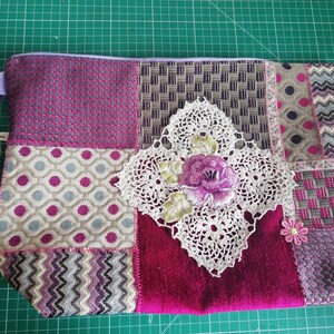 May include: A patchwork fabric zippered pouch with a purple, pink, and white color scheme. The pouch has a crocheted flower design and a purple strap.