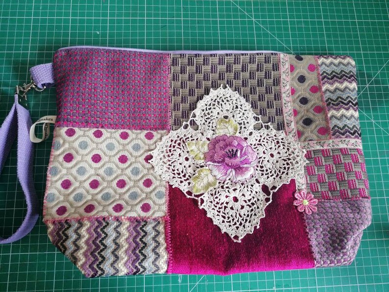 May include: A patchwork fabric zippered pouch with a purple, pink, and white color scheme. The pouch has a crocheted flower design and a purple strap.