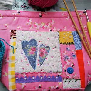 May include: Pink patchwork fabric project bag with two wooden knitting needles and a yellow crochet hook. The bag has a floral design and two blue hearts.