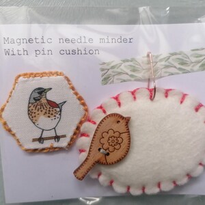 May include: A magnetic needle minder with a pin cushion. The needle minder is a small, hexagonal piece of fabric with a bird design. The pin cushion is a white oval with a red stitched edge and a wooden bird design.