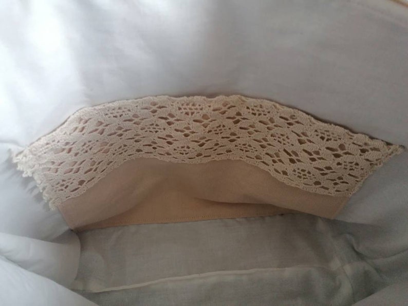 May include: A white fabric bag with a beige lace trim. The bag is open and the inside is lined with a beige fabric.