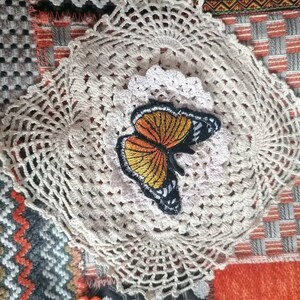 May include: A white crocheted doily with a butterfly applique in orange, yellow, and black. The doily is on a patchwork background with orange, gray, and white fabric.