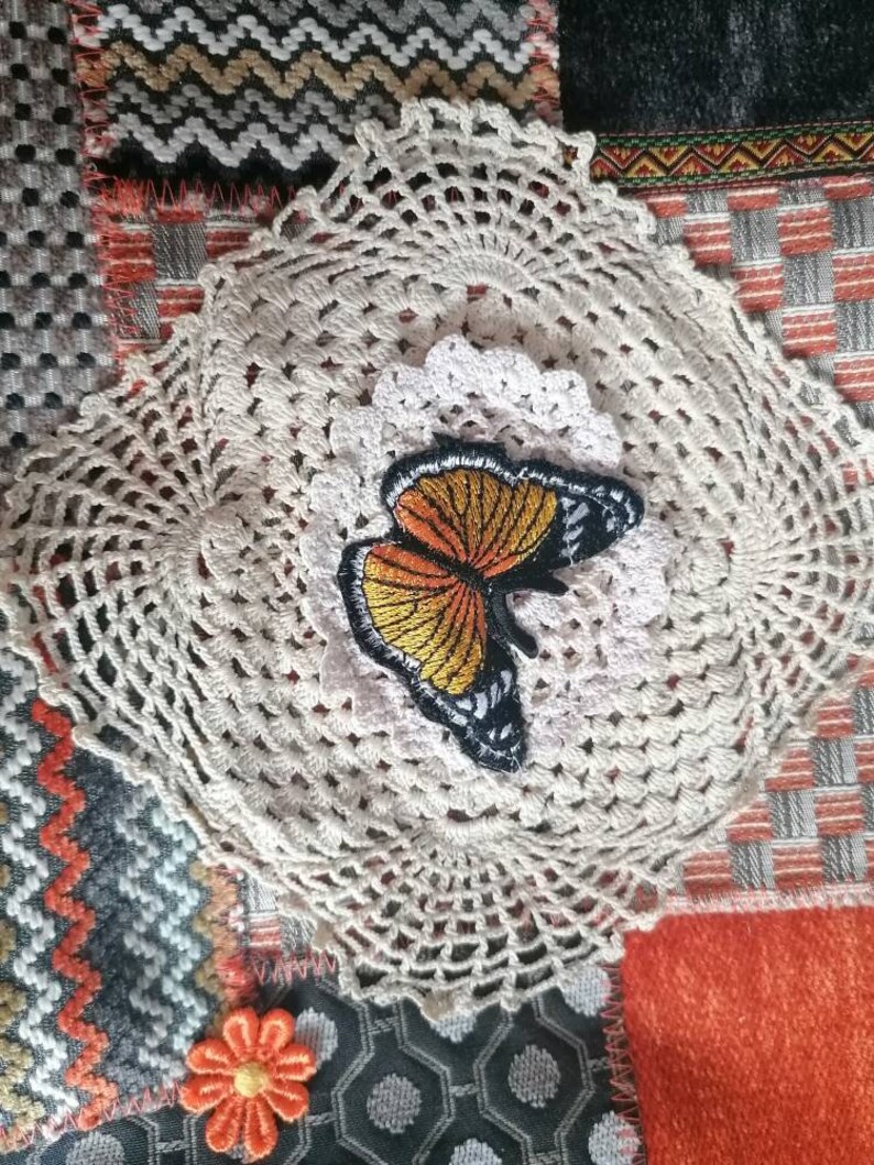 May include: A white crocheted doily with a butterfly applique in orange, yellow, and black. The doily is on a patchwork background with orange, gray, and white fabric.