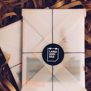 May include: A white gift box with a black circle sticker that says "LAND AND SEA" on it. The box is wrapped in white tissue paper and tied with black string.