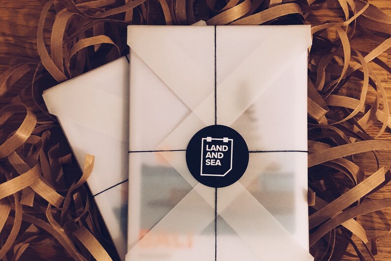May include: A white gift box with a black circle sticker that says "LAND AND SEA" on it. The box is wrapped in white tissue paper and tied with black string.
