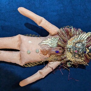 May include: A handmade doll with a pink fabric body, gold wings, and a gold lion head with a red jewel. The doll is decorated with gold and red fringe.