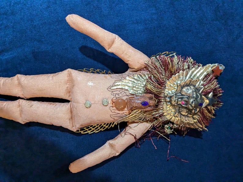 May include: A handmade doll with a pink fabric body, gold wings, and a gold lion head with a red jewel. The doll is decorated with gold and red fringe.