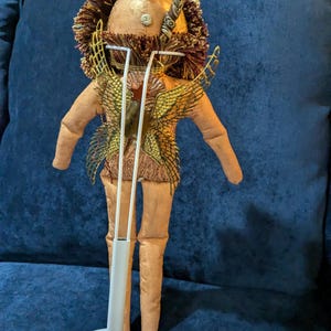 May include: A handmade doll with a gold and brown head, gold wings, and a brown body. The doll is standing on a white stand.