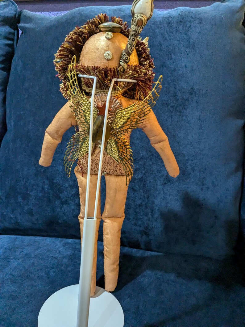 May include: A handmade doll with a gold and brown head, gold wings, and a brown body. The doll is standing on a white stand.
