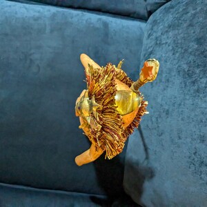 May include: A gold-colored decorative object with a lion's head and a red star on top. The object is made of metal and has a fringe of gold tassels.