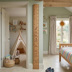 May include: A wooden growth chart with engraved markings and names, mounted on a light green wall. The chart features a rocket, flower, and other illustrations. Measurements are in inches. The room includes a teepee, toys, and a bed.