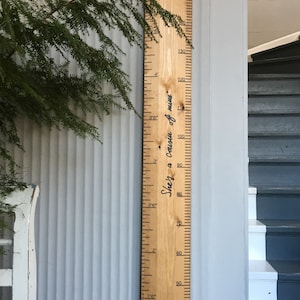 May include: A wooden growth chart with inch and foot markings, and the phrase "She's a cousin of mine" in cursive. The chart is light brown and mounted on a light gray wall. Green foliage is visible on the left side of the image.