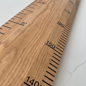 May include: Wooden height chart with black markings and numbers. The chart measures up to 6 feet, with markings in inches and feet. The wood has a natural grain and a light brown color. The numbers are in a bold, sans-serif font.
