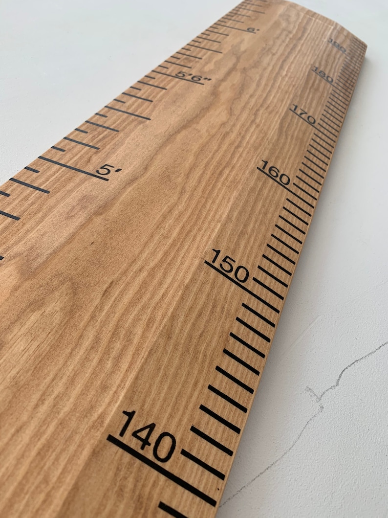 May include: Wooden height chart with black markings and numbers. The chart measures up to 6 feet, with markings in inches and feet. The wood has a natural grain and a light brown color. The numbers are in a bold, sans-serif font.