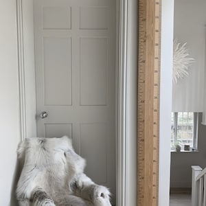 May include: A wooden growth chart with black markings, measuring up to 6 feet 3 inches. The chart is mounted on a white wall next to a white door and a chair draped with a faux fur throw. The room has a neutral color palette.