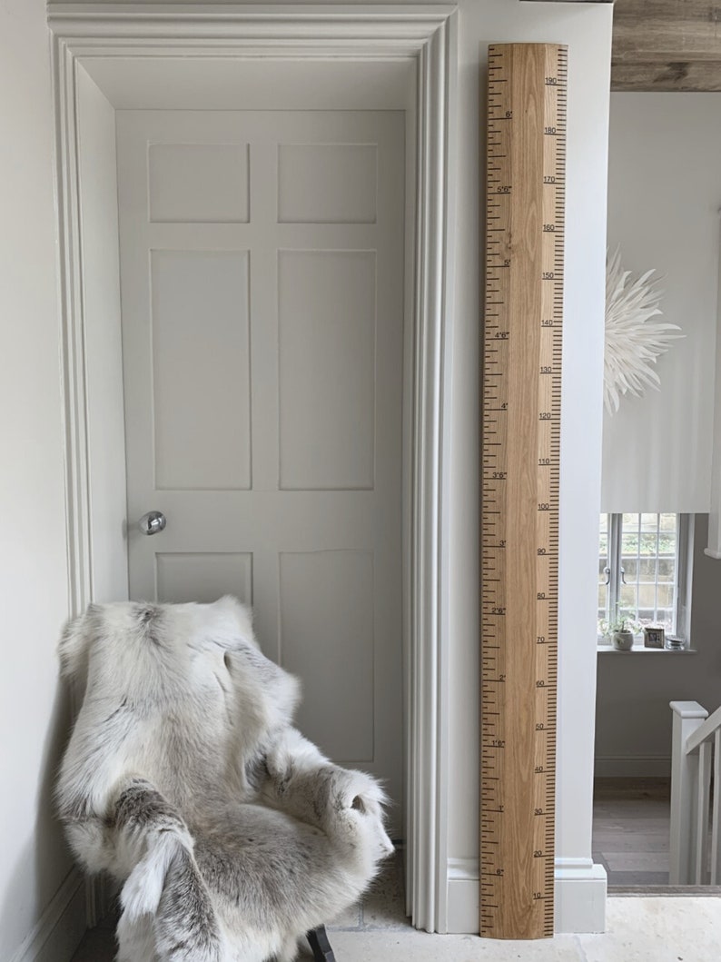 May include: A wooden growth chart with black markings, measuring up to 6 feet 3 inches. The chart is mounted on a white wall next to a white door and a chair draped with a faux fur throw. The room has a neutral color palette.