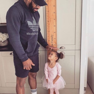May include: A wooden height chart with measurements in inches is held by a person, measuring a young child. The child is wearing a pink leotard and tutu. The height chart has the words "The [Family]" printed on it.