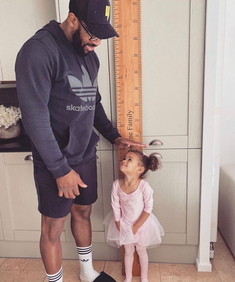 May include: A wooden height chart with measurements in inches is held by a person, measuring a young child. The child is wearing a pink leotard and tutu. The height chart has the words "The [Family]" printed on it.