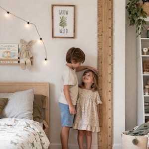 May include: A wooden growth chart with inch markings, next to two children. The chart is mounted on a white wall, with a framed print that reads "Grow Wild". The children are standing next to the chart, one measuring the other's height.