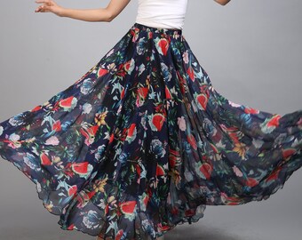 May include: A woman wearing a white tank top and a long, flowing floral skirt. The skirt is navy blue with a colorful floral print that includes red, pink, and green flowers. The skirt is gathered at the waist and has a flowing, A-line silhouette.