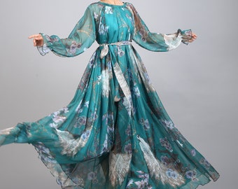 Puede incluir: Un vestido largo de color turquesa con estampado floral y de plumas de pavo real. El vestido tiene mangas largas y transparentes y un cinturón a juego. La modelo lleva tacones blancos. El vestido es de tejido ligero y fluido.