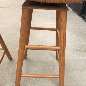 May include: A wooden bar stool with a woven wicker seat. The stool has a simple design with a rectangular seat and four legs.