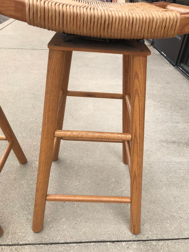 May include: A wooden bar stool with a woven wicker seat. The stool has a simple design with a rectangular seat and four legs.