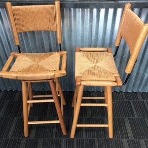 May include: Two wooden bar stools with woven cane seats and backs. The stools have a black metal frame and are made of natural wood.