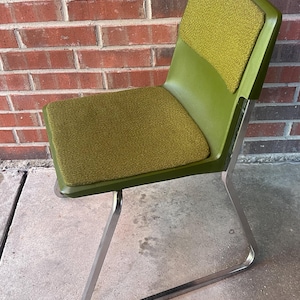 May include: A vintage green chair with a curved back and seat. The seat and backrest have a textured, olive-green fabric. The chair's frame is made of polished metal. The chair is set against a red brick wall.