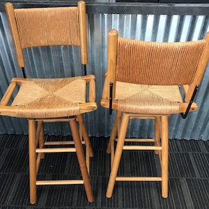 May include: Two wooden bar stools with woven cane seats and backs. The stools have a natural wood finish and black metal accents.