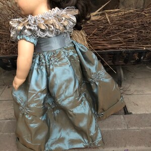 May include: A teal and brown doll dress with a silver and gold beaded collar. The dress has a ruffled skirt and a fitted bodice.