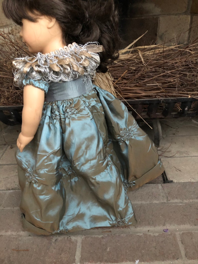 May include: A teal and brown doll dress with a silver and gold beaded collar. The dress has a ruffled skirt and a fitted bodice.